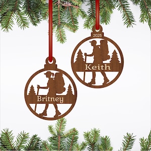 Può includere: Due decorazioni natalizie in legno con nastri rossi. Ogni ornamento presenta una silhouette di escursionista, alberi e un nome. Uno ha il nome "Britney", l'altro "Keith" e l'anno "2025".