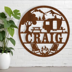Pode incluir: Um sinal redondo de madeira marrom com um trailer, montanhas, árvores, uma fogueira e duas cadeiras. O nome "CRAIG" está escrito em letras grandes abaixo do trailer.