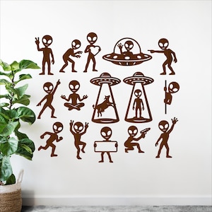 May include: Brown alien wall decals in various poses, including peace signs, holding hearts, and flying in UFOs. One alien is abducting a cow. The decals are arranged on a white wall with a potted plant in the corner.