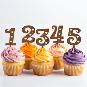 May include: Five cupcakes decorated with wooden number cake toppers, 1, 2, 3, 4, and 5. The cupcakes are frosted in pink, orange, yellow, peach, and purple.