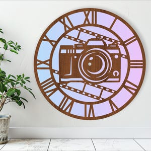 Puede incluir: Un reloj de pared de madera con un diseño de cámara y números romanos. La esfera del reloj tiene un degradado de azul claro a rosa.