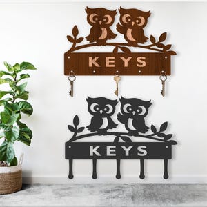 May include: Two owl-themed key holders. The top one is brown with the word "KEYS" and three key hooks. The bottom one is black with the word "KEYS" and four key hooks. Both have owl silhouettes perched on branches.