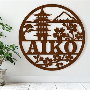 May include: A brown wooden wall hanging with a circular design featuring a Japanese pagoda, mountain, cherry blossoms, and the name "AIKO".