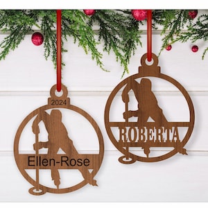 May include: Two wooden hockey player ornaments with personalized names. The ornament on the left says "Ellen-Rose" and the ornament on the right says "Roberta".