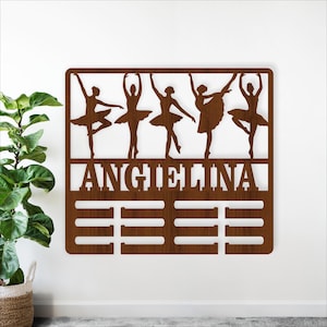 May include: A wooden wall hanging featuring five ballerina silhouettes in various poses. The name "ANGELINA" is cut out below the dancers, with three horizontal bars beneath the name. The piece is made of brown wood and is mounted on a white wall.