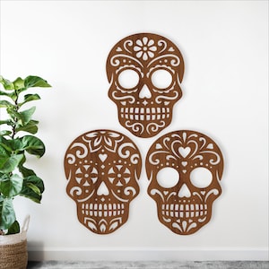 Peut inclure: Trois décorations murales en bois représentant des têtes de mort mexicaines avec des motifs découpés complexes. Les crânes sont bruns avec des motifs floraux et en forme de cœur, et sont montés sur un mur blanc. Thème Dia de los Muertos.