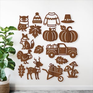 May include: Wooden cutouts of fall-themed items, including an owl, fox, turkey, deer, pumpkins, leaves, a truck, a wheelbarrow, a sweater, a hat, a beanie, and mushrooms. The cutouts are brown and arranged on a white wall.