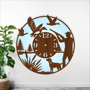 May include: A round, wooden wall clock with a hunting theme. The clock features a hunter, dog, ducks, trees, and reeds. The clock face has the numbers 12, 3, 6, and 9. The background is light blue.