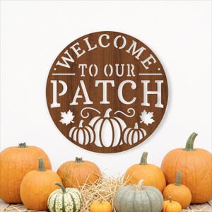 May include: A circular brown wood-toned sign featuring white cut-out text that reads "WELCOME TO OUR PATCH" with a graphic of three pumpkins and two maple leaves centered below the text, displayed above a collection of various orange and green pumpkins.