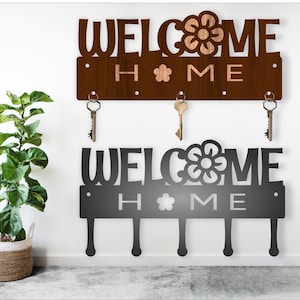 May include: Two key holders with the words "WELCOME HOME" cut out. One is brown with a flower design, the other is black. Both have key hooks. A potted plant is in the background.