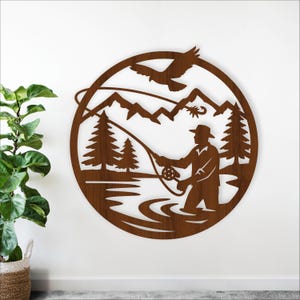 May include: A brown metal wall art piece depicting a fisherman fly fishing in a river, with mountains, trees, and a soaring bird in the background. The artwork is circular and has a rustic, outdoorsy theme.