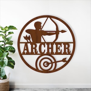 Pode incluir: Uma peça de arte de parede circular de metal castanho com um arqueiro com arco e flecha, um alvo e a palavra "ARCHER". O arqueiro está no centro do círculo, apontando para um alvo com uma flecha.
