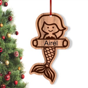 May include: Wooden Christmas ornament featuring a mermaid with a tail and the name "Aariel" on a banner.