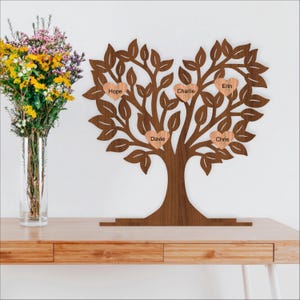 Op de afbeelding: Een houten familiestamboom met een hartvormige kroon en een stevige stam. Vijf hartvormige naamlabels zijn aan de takken bevestigd, elk met een naam. De boom staat op een houten tafel, met een vaas bloemen links.