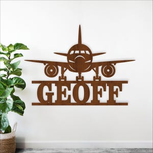 May include: A brown metal airplane wall art piece with the name "GEOFF" cut out below the plane. The airplane has wings, a tail, and visible engines. The art is mounted on a white wall.
