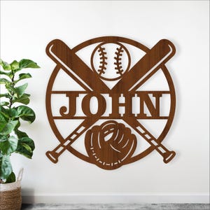 May include: A brown wooden baseball-themed wall sign featuring the name "JOHN". The design includes a baseball, crossed bats, and a baseball glove within a circular frame. The sign is mounted on a white wall. The sign is likely made of wood.