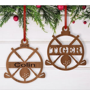 May include: Two wooden Christmas ornaments shaped like golf balls with crossed golf clubs. One ornament has the name "Colin" and the year "2024" on it. The other ornament has the name "Tiger" on it.
