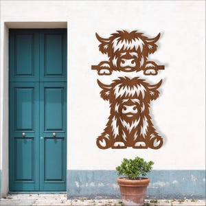 May include: Two wooden Highland cow wall art pieces. The top cow is peeking over a horizontal bar, while the bottom cow is sitting. Both have shaggy fur and large horns. The art is a warm brown colour and mounted on a white wall.