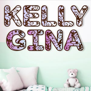 May include: A decorative wall art piece with the names "KELLY GINA" spelled out in large, stylized letters. The letters are decorated with a purple and pink gradient pattern and feature a fairy silhouette design.