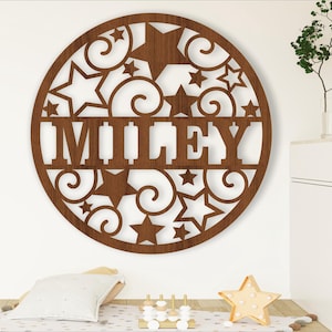 Könnte beinhalten: Ein rundes Schild aus braunem Holz mit einem ausgeschnittenen Design aus Sternen und Wirbeln. Der Name "MILEY" ist in der Mitte des Schildes ausgeschnitten.