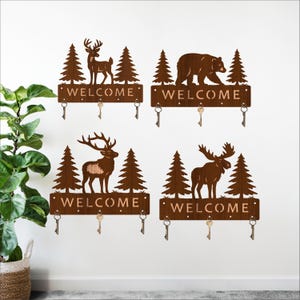 May include: Four rustic metal key holders with a nature theme. Each features a different animal silhouette: a deer, a bear, and a moose, along with the word "WELCOME". Each holder has four key hooks. The key holders are mounted on a white wall.