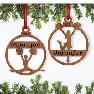 May include: Two wooden Christmas baubles shaped like a circle with a cheerleader silhouette inside. The baubles are personalised with the names "Monique" and "Jennifer" and the year "2024".