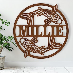 Puede incluir: Un letrero redondo de madera con dos peces estilizados y el nombre "Millie" en una fuente decorativa.