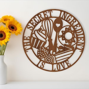 May include: A brown wooden sign with the text "The Secret Ingredient is Love" in a white circle. The circle is decorated with a whisk, a heart-shaped spoon, a cupcake, and other baking-themed designs.