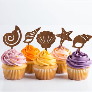 Puede incluir: Seis cupcakes decorados con adornos de madera en forma de conchas marinas, una estrella de mar y una espiral. Los cupcakes están cubiertos de glaseado rosa, naranja, amarillo y morado.