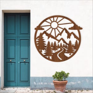 May include: A round, brown wooden wall art piece featuring a mountain and forest scene. The design includes a sun, mountains, trees, clouds, and a winding road. The artwork is mounted on a white wall near a teal door and a potted plant.
