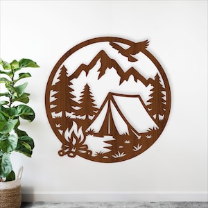 May include: A circular wooden wall art piece featuring a camping scene. The design includes mountains, pine trees, a tent, a campfire, and a flying bird. The artwork is cut from brown wood and is mounted on a white wall.
