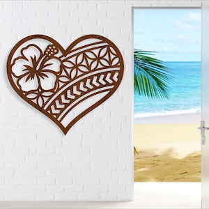 Op de afbeelding: Bruine houten hartvormige wanddecoratie met een uitgesneden hibiscusbloem en geometrische patronen. Het hart is gemonteerd op een witte bakstenen muur, met uitzicht op een strand en de oceaan.