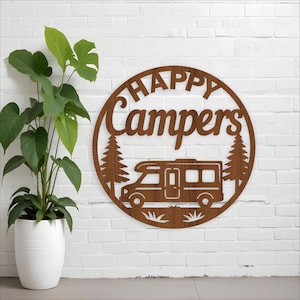 May include: A circular, brown wooden sign with the words "HAPPY Campers" cut out. The sign depicts a camper van, two trees, and grass. It's mounted on a white brick wall, with a potted plant to the left.