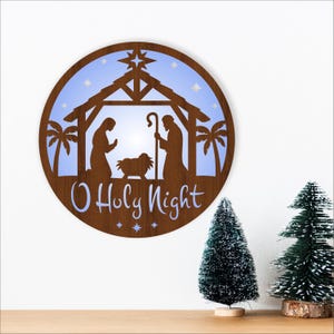 May include: A round, wooden Christmas decoration featuring a nativity scene silhouette with the text "O Holy Night." The design includes a stable, palm trees, stars, and figures of Mary, Joseph, and baby Jesus. The background is light blue.