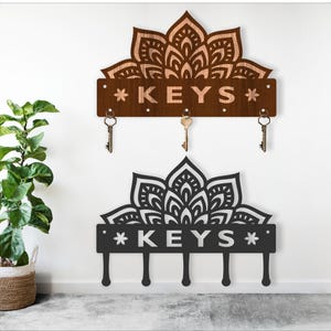 May include: Two key holders with a mandala design. One is brown with the word "KEYS" and three key hooks. The other is black with the word "KEYS" and four key hooks. Both are mounted on a white wall.