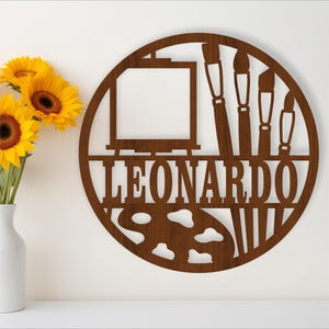 May include: A brown wooden round sign with a paint palette, paintbrushes, and an easel. The name "LEONARDO" is written in a decorative font below the image.