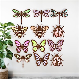 May include: A collection of wooden insect cutouts, including dragonflies, bees, butterflies, and ladybugs. The insects are in various colors, such as green, pink, and purple, and are mounted on a white wall. The cutouts are a decorative element.
