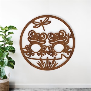 May include: A round, wooden wall art piece featuring two cartoon frogs and a dragonfly. The frogs are brown with large eyes and are sitting on a log. The dragonfly is above them. The background is white.