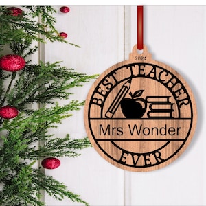 Może przedstawiać: Okrągła drewniana ozdoba z tekstem "Best Teacher Ever" i "Mrs. Wonder" w czarnym k&oacute;łku. Ozdoba zawiera r&oacute;wnież oł&oacute;wek, jabłko i książki.