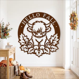 May include: A circular dark brown wooden wall sign featuring a cutout illustration of a Highland cow surrounded by oak leaves. The text HELLO FALL is engraved along the top curve of the circular frame, creating a decorative seasonal wall hanging.