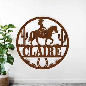 May include: A circular, brown metal wall art piece featuring a silhouette of a cowgirl riding a horse, with cacti on either side. The name "CLAIRE" is displayed below the horse, with a horseshoe at the bottom. The design is cut out of the metal.