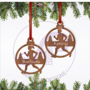 Puede incluir: Dos adornos navideños de madera con cintas rojas. Cada adorno presenta un recorte de una persona corriendo, con los nombres "Barbara" y "Kenny" inscritos. Los adornos están decorados con pinos y el año 2025.