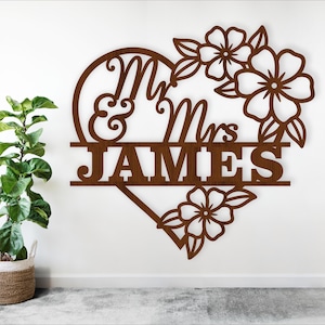 May include: A brown wooden sign with the words "Mr & Mrs JAMES" cut out in a heart shape, adorned with floral accents. The sign is displayed on a white wall, with a potted plant in the foreground.