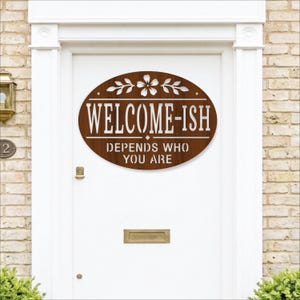 Pode incluir: Uma placa oval de madeira castanha com um design floral branco no topo e o texto branco "WELCOME-ISH" seguido de "DEPENDS WHO YOU ARE" numa fonte estilo stencil, montada numa porta branca com uma ranhura para correio dourada.