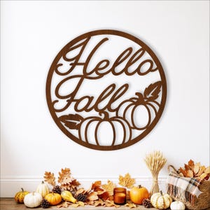 May include: A circular brown wooden wall sign featuring the cutout text Hello Fall in a cursive script. The design includes two stylized pumpkins and leaves integrated into the lower portion of the circular frame, creating a decorative seasonal piece.