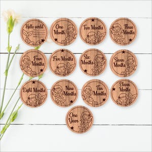 May include: Wooden mermaid milestone discs for baby's first year. Each disc is round with a mermaid illustration and month labels from "One Month" to "One Year". The "Hello World" disc has space for baby's name and birth details. A charming baby shower gift.