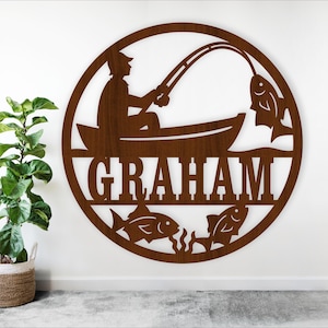 Pode incluir: Uma peça de arte de parede circular de metal castanho com um pescador num barco, uma cana de pesca e peixes. O nome "GRAHAM" é recortado no centro. Decorativo e adequado para uma casa ou cabana.
