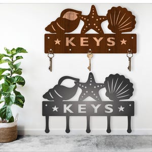 May include: A wooden key holder with a seashell, starfish, and the word "KEYS" in a brown wood finish. The key holder has five hooks for hanging keys.