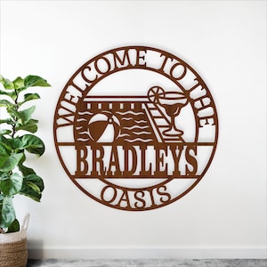 Könnte beinhalten: Ein runder, brauner Metallschild mit den Worten "WELCOME TO THE" oben und "BRADLEYS OASIS" unten. In der Mitte befindet sich eine Poolszene mit einem Strandball, einem Pool, einer Leiter und einem Cocktail. Das Schild ist an einer weißen Wand befestigt.