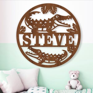 Op de afbeelding: Een rond bord van bruin hout met een uitgesneden ontwerp van twee alligators en de naam "STEVE" in het midden. Het bord is perfect voor een kinderkamer of een crèche.